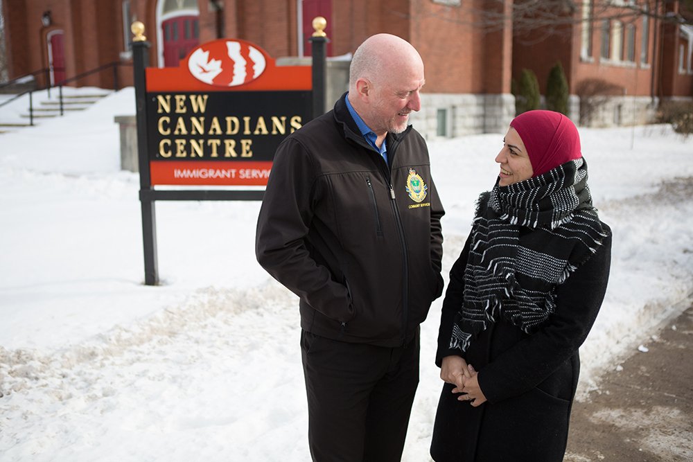 2 individuals stand in front of the New Canadians Centre having a jovial conversation