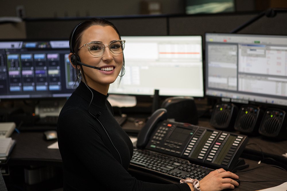 A woman smiles at a camera with a head set on her heat in front of a dispatch desk with multiple computers, speaks and phones sit behind her