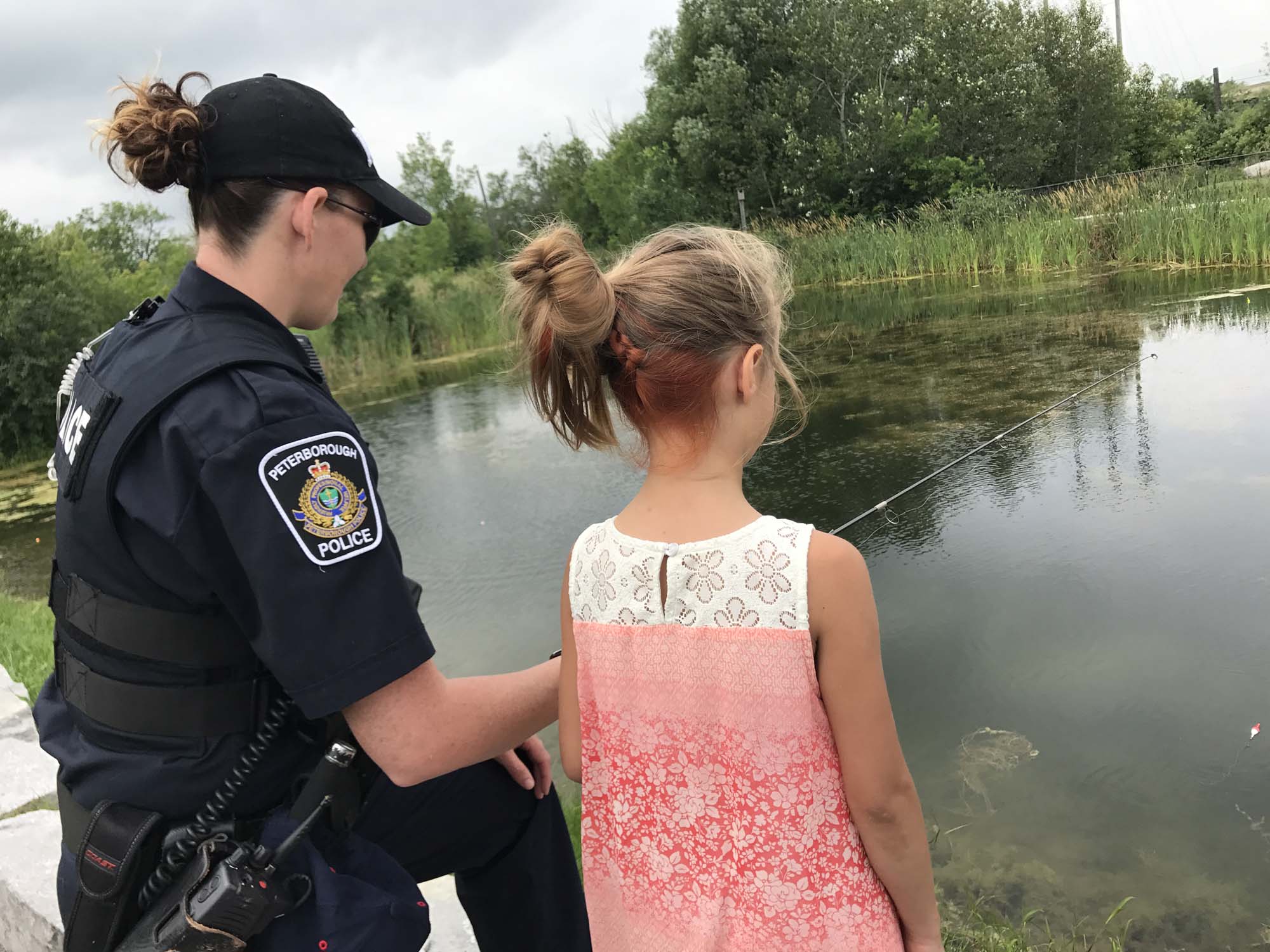 A Peterborough police officer stands with a little girl fishing in a pond