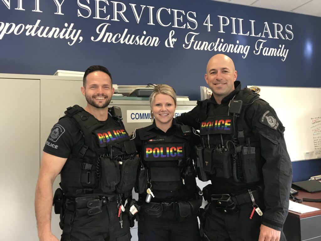 3 Police officers stand together with their arms wrapped around each other with Pride flag coloured Police designation on their chests